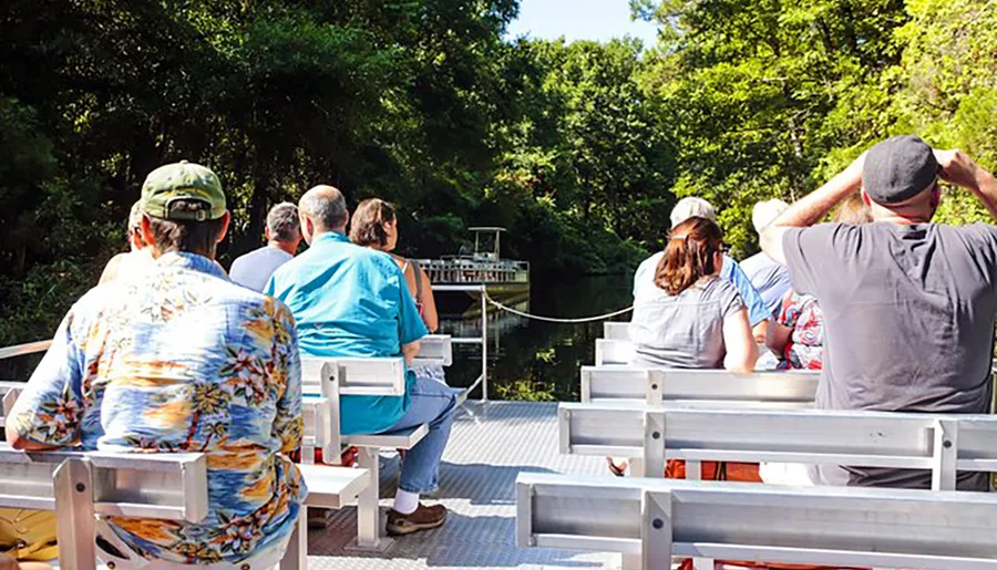 A group of people are seated on a boat, looking out towards a lush green riverbank on a sunny day.
