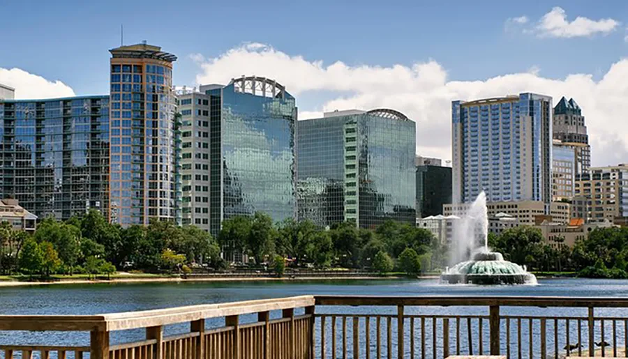 The image shows a tranquil urban park with a lake featuring a fountain in the foreground and a backdrop of modern skyscrapers under a clear sky.