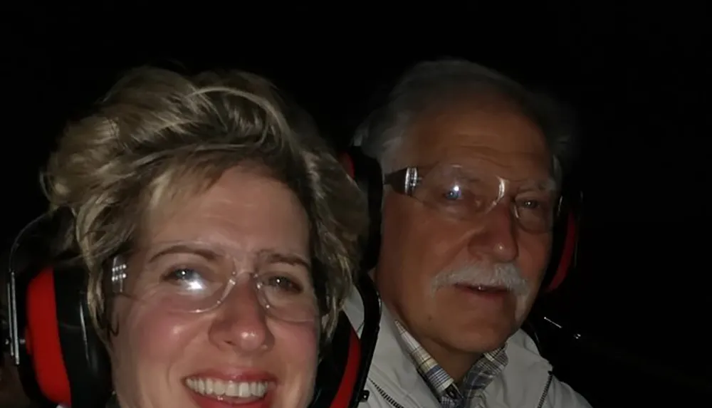 A woman and a man are wearing red earmuffs in a dimly lit setting with the woman smiling at the camera and the man looking on more seriously