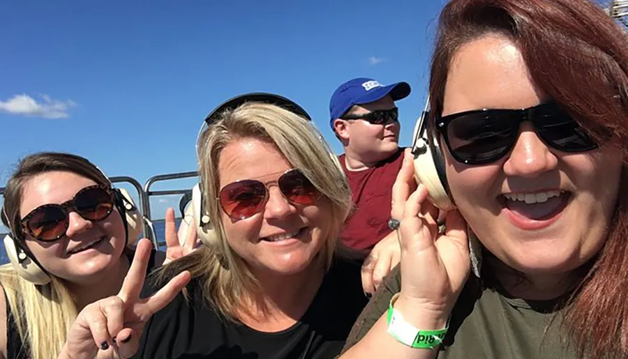 Three people are smiling and taking a selfie while wearing headphones, possibly on a tour or an open vehicle, under a clear blue sky.