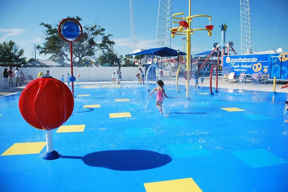 Children play in a colorful sunlit water park with various sprinklers and water features on a clear day