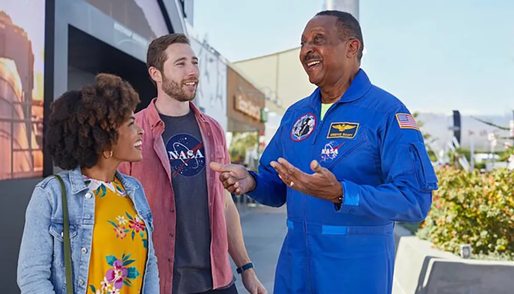 A person in a NASA flight suit is engaging in conversation with two individuals who appear fascinated by the discussion set against a background that suggests a space-related exhibition or environment