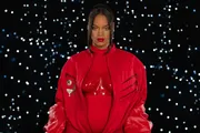 The image shows a person wearing a glossy red outfit with matching lipstick, posing confidently against a backdrop dotted with numerous small white lights.