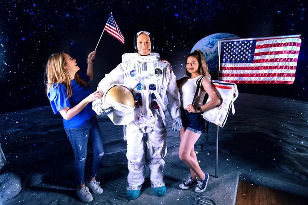 Two people are posing playfully with a spacesuit-clad mannequin against a space-themed backdrop complete with a lunar surface an American flag and the Earth in the distance