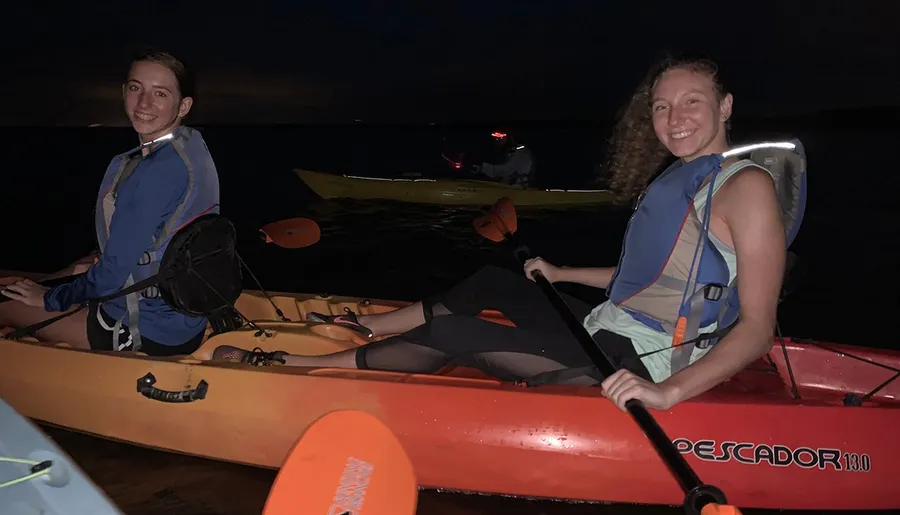 Two people are smiling and seated in kayaks, wearing life vests at night with another kayak visible in the background.