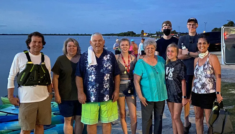 A group of eight people of various ages pose together for a photo near a waterfront at dusk, with some wearing life vests and sporting a relaxed, outdoor outing attire.