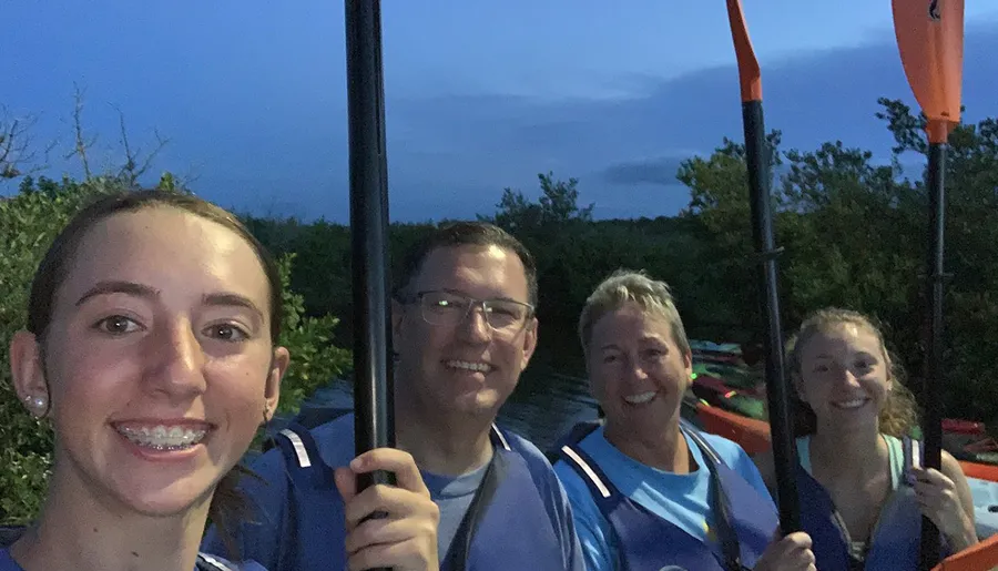 Four people are smiling for a group selfie during twilight, holding kayak paddles and wearing life jackets, suggesting they are about to embark on or have returned from a kayaking adventure.