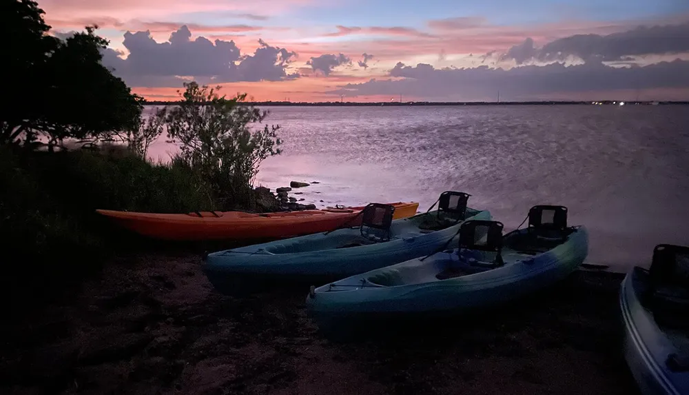 A tranquil scene showing several kayaks resting on a beach at dusk with a colorful sunset over the water