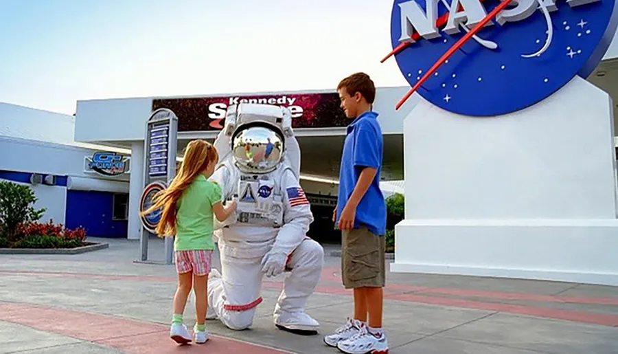 A person in an astronaut suit is kneeling to interact with a young girl while a boy stands nearby, in front of the Kennedy Space Center sign.