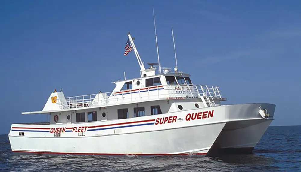 A white and red boat named SUPER QUEEN is cruising on blue water under a clear sky displaying an American flag at its stern