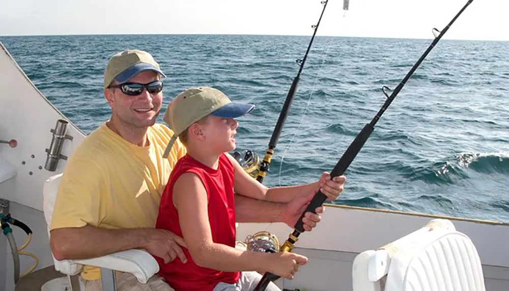 A smiling man in sunglasses and yellow attire guides a young boy in a red sleeveless shirt and cap as they fish together on a boat at sea