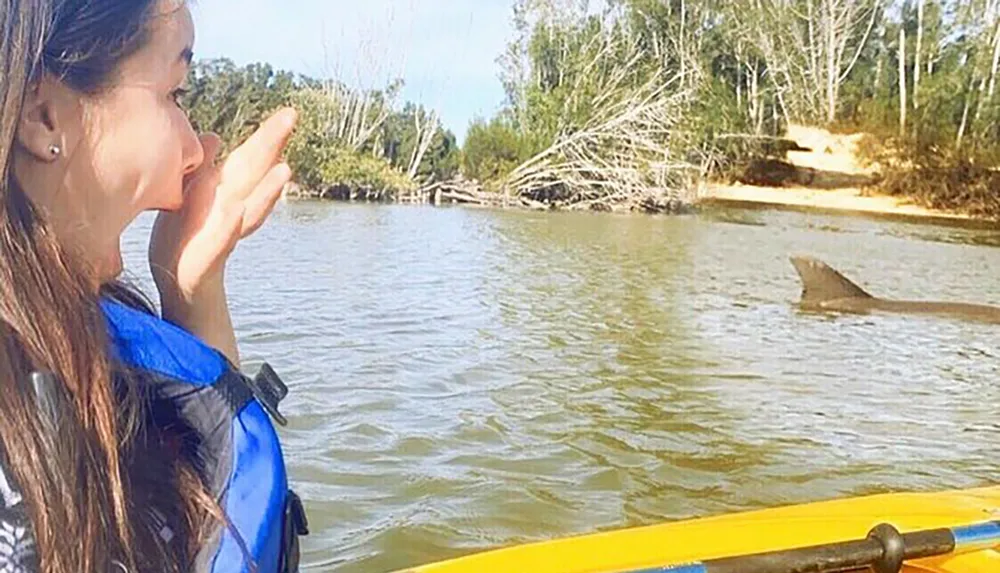 A person is seemingly blowing a kiss to a shark fin above the water while kayaking