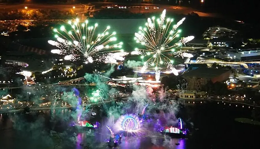 This is an aerial night-time view of a festive fireworks display over a brightly illuminated park with a Ferris wheel.