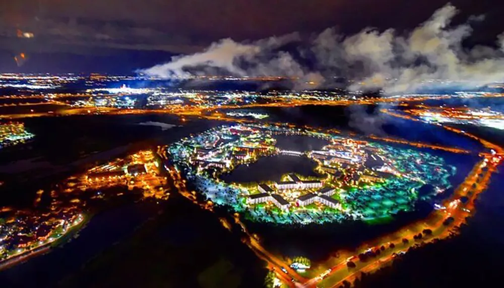 The image shows a vibrant aerial nighttime view of a brightly lit complex with adjacent roads and surrounding dark landscape under a cloudy sky