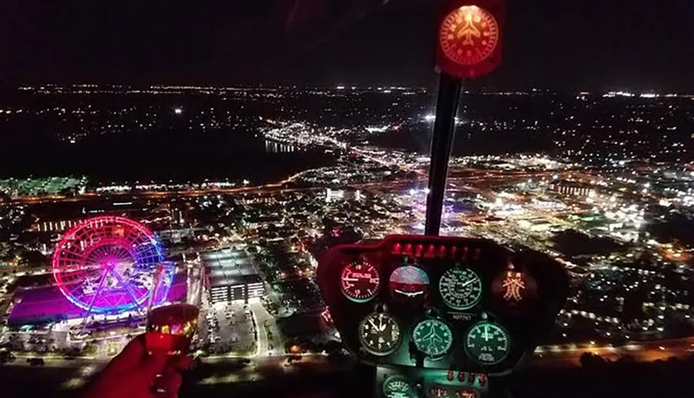 A helicopter cockpit view at night shows a vibrant illuminated cityscape with a Ferris wheel prominently displayed
