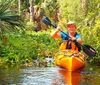 Two people are smiling and posing with peace signs while kayaking together in a verdant river setting