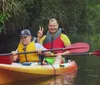 Two people are smiling and posing with peace signs while kayaking together in a verdant river setting
