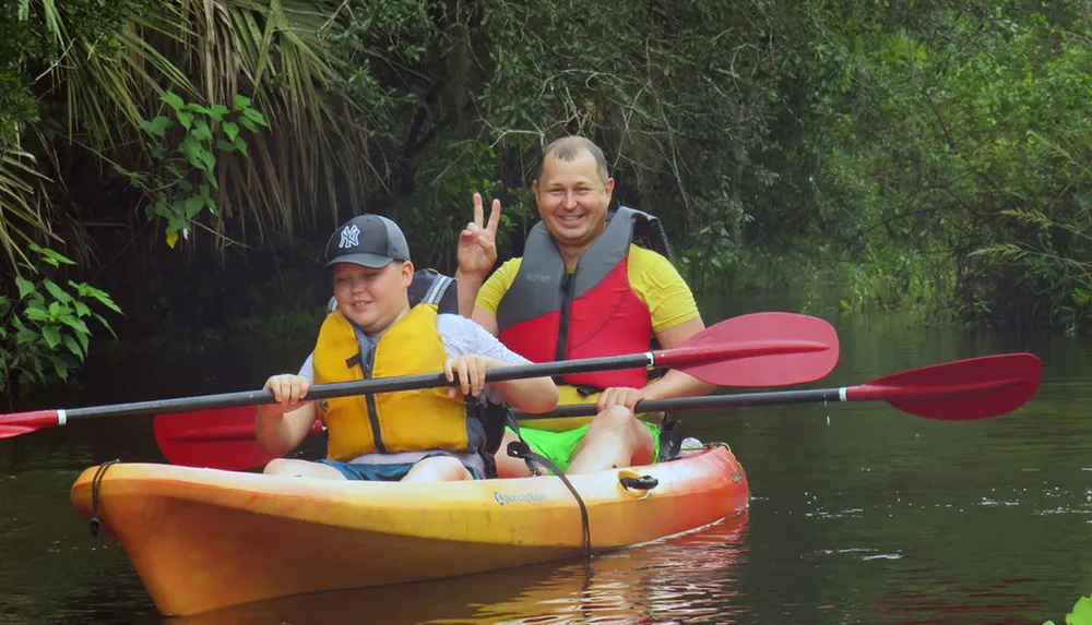 Two people are smiling and posing with peace signs while kayaking together in a verdant river setting