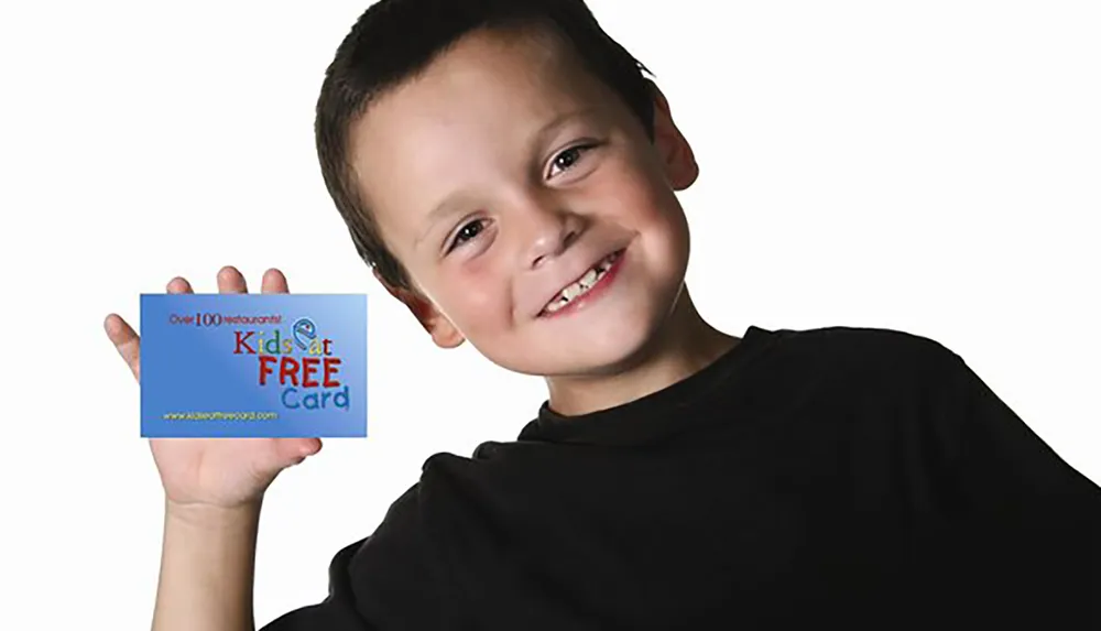 A cheerful young boy is holding up a blue card that promotes some sort of Kids Eat Free offer