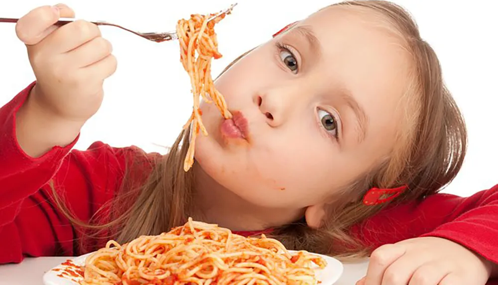 A child is making an amusing face while attempting to eat a messy plate of spaghetti with her fork