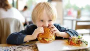 A young child is taking a big bite out of a hamburger at a restaurant table with a salad on the side.