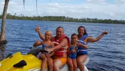 A family of four is posing with thumbs up on a yellow jet ski in a large body of water, surrounded by trees under a partly cloudy sky.