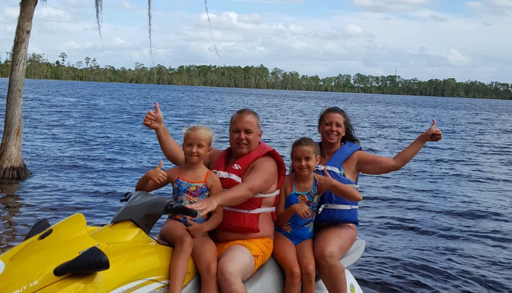 A family of four is posing with thumbs up on a yellow jet ski in a large body of water surrounded by trees under a partly cloudy sky