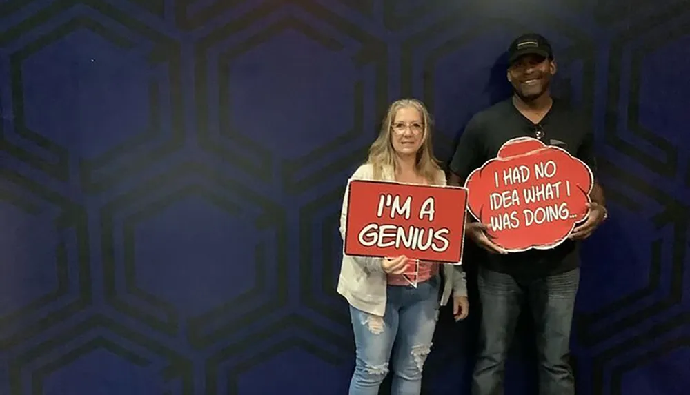 A woman and a man are posing with humorous signs one reading IM A GENIUS and the other I HAD NO IDEA WHAT I WAS DOING against a patterned blue backdrop