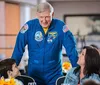 An individual in a NASA astronaut suit is leaning over a table to interact with smiling people seated around it