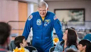 An individual in a NASA astronaut suit is leaning over a table to interact with smiling people seated around it.