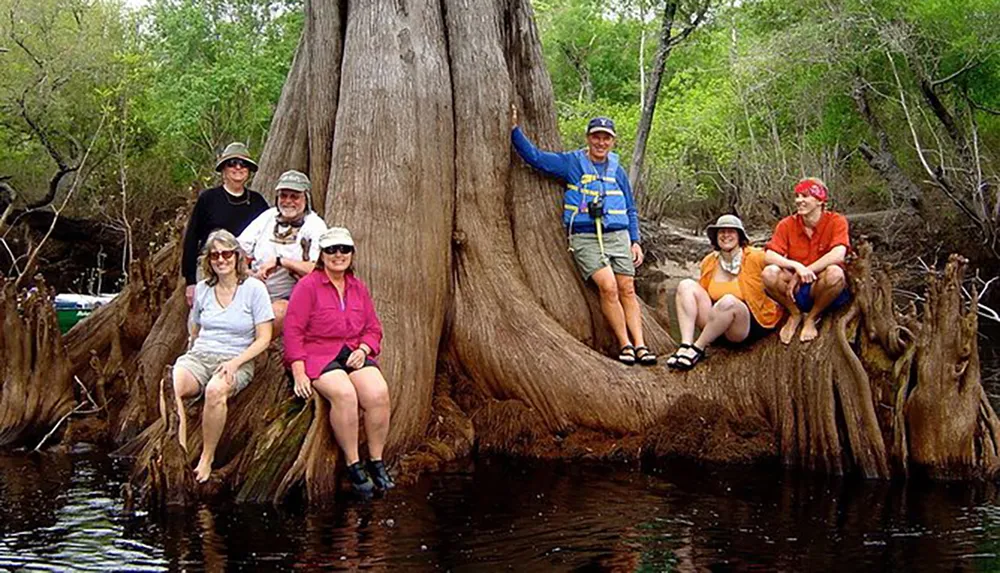 A group of people in outdoor attire are posing cheerfully around a large tree with protruding roots by a body of water