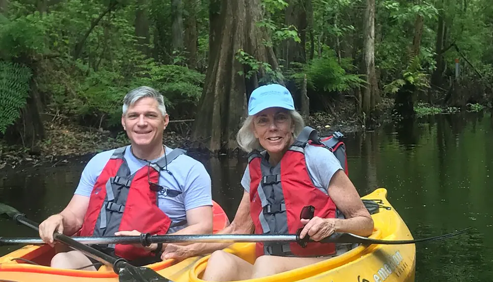 Two people are smiling while kayaking together on a calm river surrounded by greenery