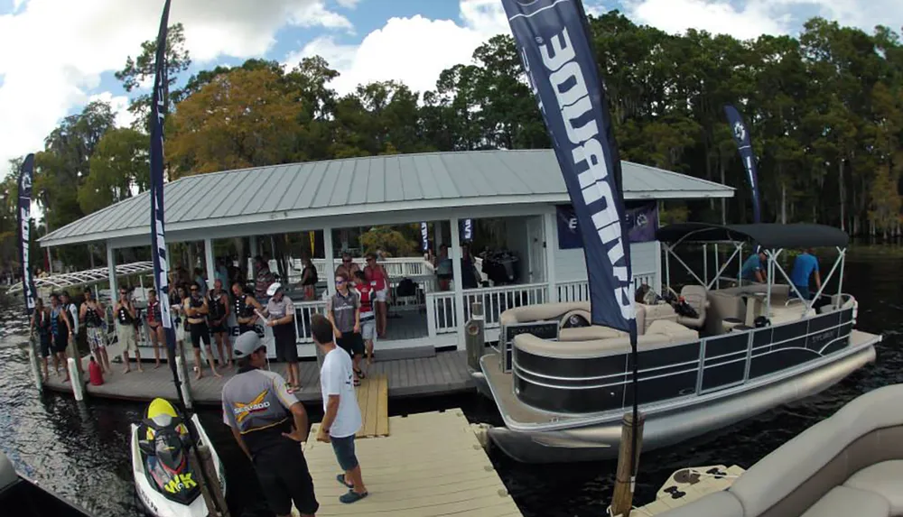 A group of people gathered on a dock by a waterway with pontoon boats nearby and promotional flags fluttering in the breeze