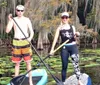 A man and a woman are standing on paddleboards in a calm body of water with lily pads surrounded by trees draped with Spanish moss