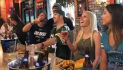 A group of friends are enjoying a lively moment at a sports bar, with some dressed in sports team apparel, sharing food, drinks, and laughter.