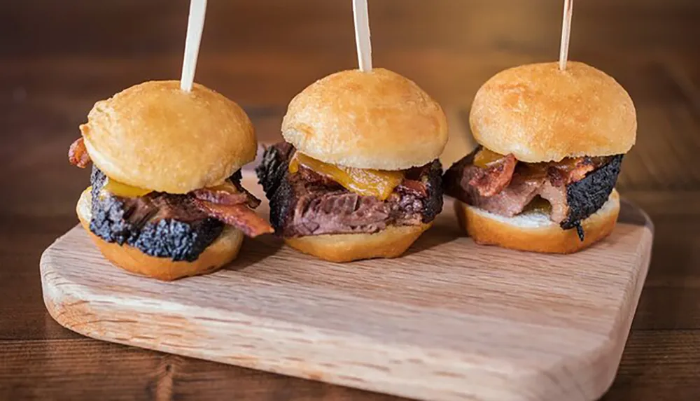 Three mini burgers with bacon and cheese are presented on a wooden cutting board