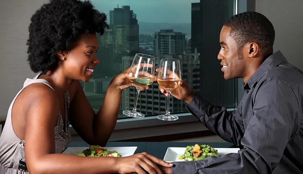 A couple enjoys a romantic dinner with wine by a window overlooking a city skyline