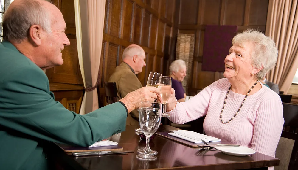 Two senior individuals are joyfully toasting with glasses of water at an elegantly set table in a dining establishment