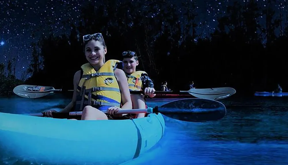 Two people are smiling while kayaking at night illuminated by a bio-luminescent glow under the water
