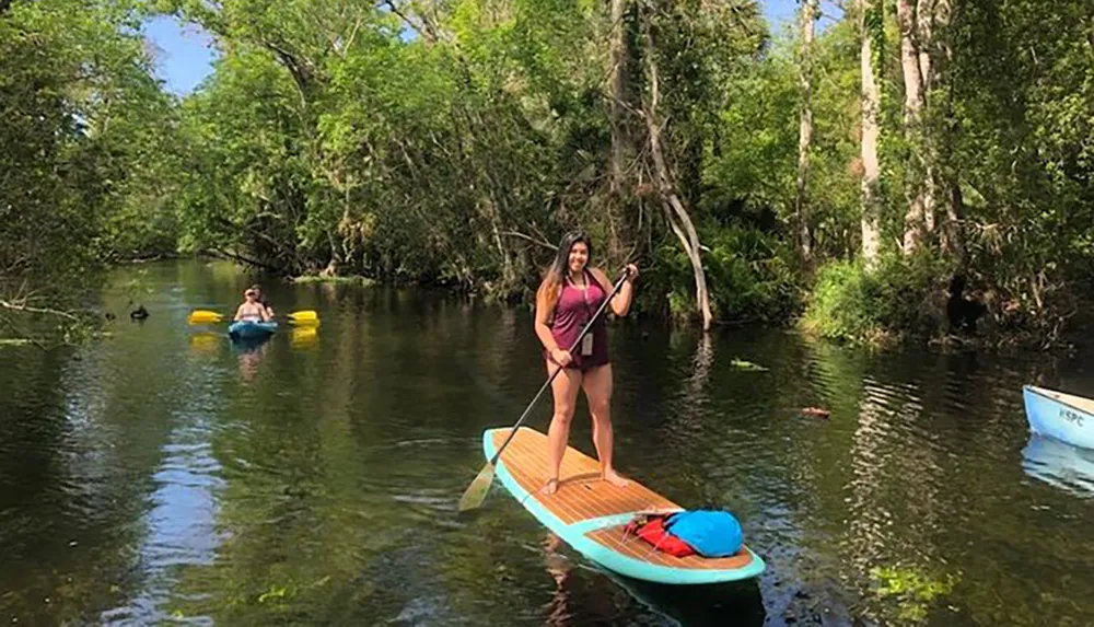 A person is standing on a paddle board navigating a calm and verdant river with others in kayaks or canoes in the background