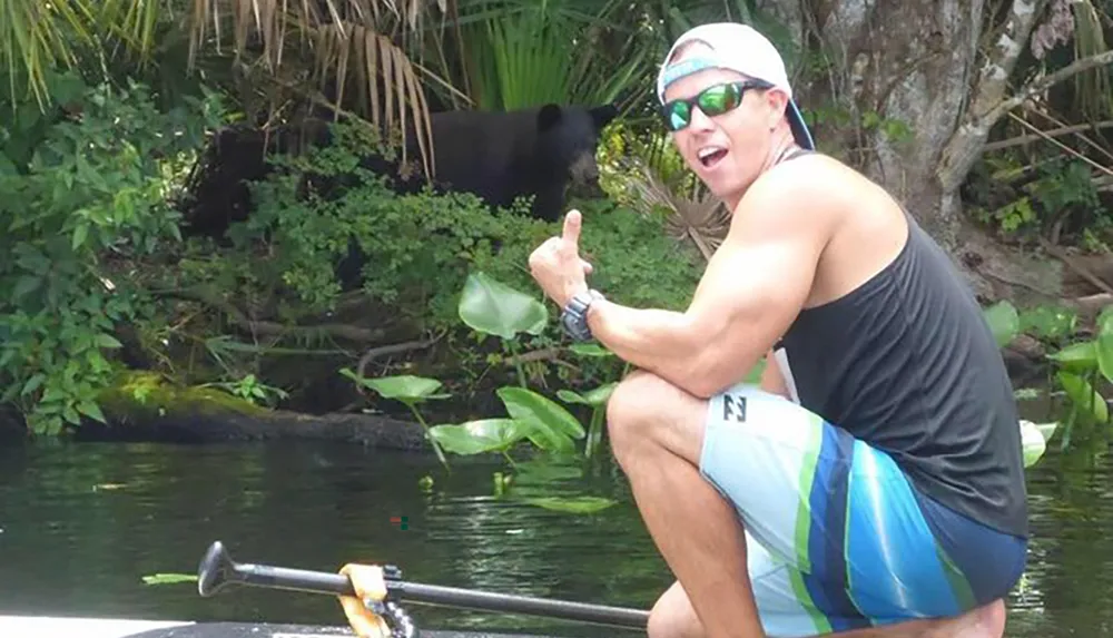 A man is giving a thumbs-up while sitting on a boat seemingly unaware of a large black animal possibly a bear in the background among the foliage