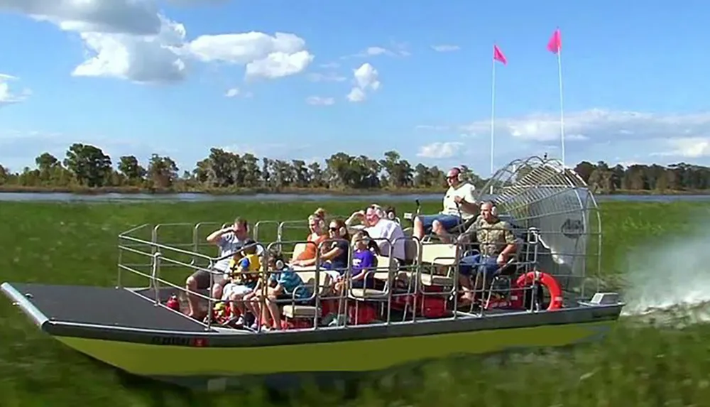 An airboat filled with passengers glides across what appears to be a marsh or wetland area with the pilot standing at the rear