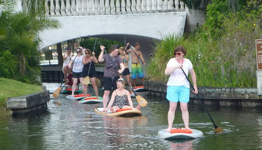 A group of people are stand-up paddleboarding on a waterway, with a bridge in the background.