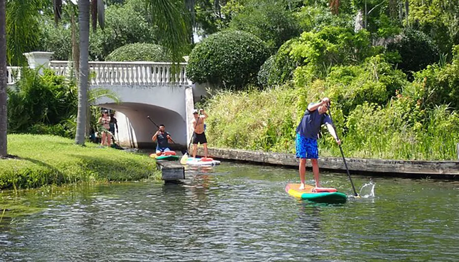 Three individuals are stand-up paddleboarding on a calm waterway under a sunny sky, passing under a white arched bridge surrounded by lush greenery.