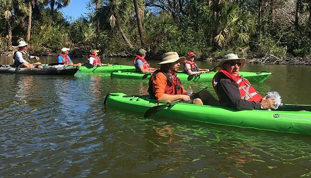 A group of people wearing life jackets and hats are paddling green kayaks down a calm river surrounded by lush vegetation