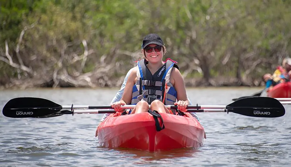 A smiling person is kayaking wearing a life jacket and sunglasses holding a double-bladed paddle with natural scenery in the background