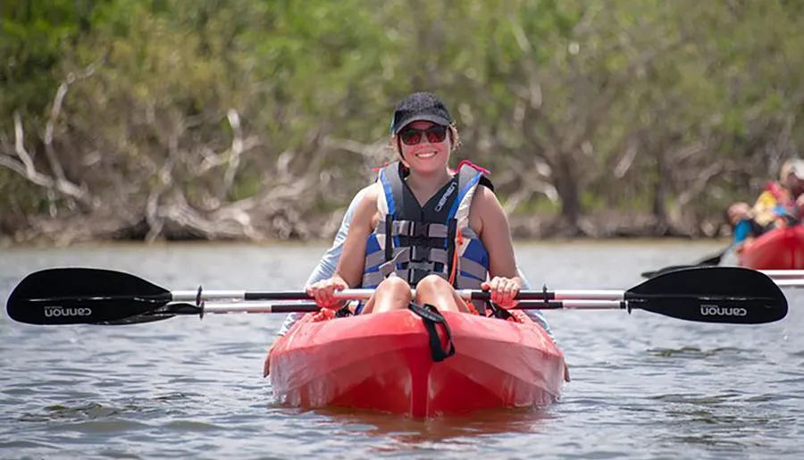A smiling person is kayaking, wearing a life jacket and sunglasses, holding a double-bladed paddle, with natural scenery in the background.