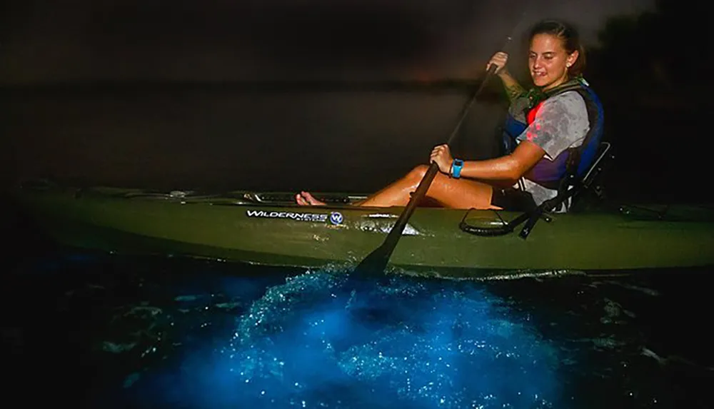 A woman is kayaking at night on water that is illuminated with a striking bioluminescent glow