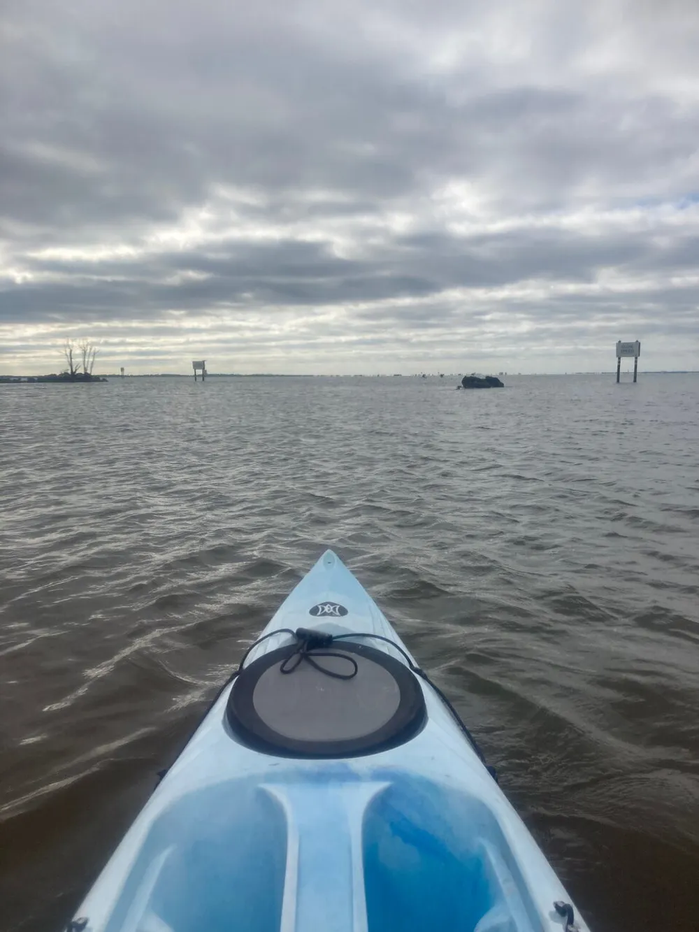 A person is kayaking on a cloudy day with the bow of their blue kayak pointing towards a distant shoreline and navigational signs over open water