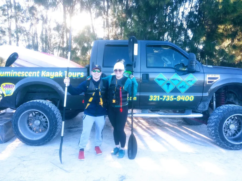 Two people are posing with paddles in front of a truck advertising Bioluminescent Kayak Tours suggesting they are about to engage in an outdoor kayaking adventure